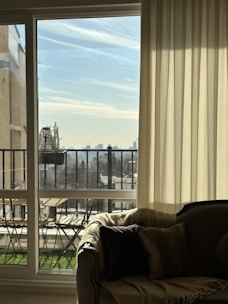 A cozy interior setting features a comfortable couch with soft pillows and a blanket, positioned next to a large window. The window is partially covered by sheer curtains, revealing an outdoor view with a balcony equipped with a small table and chairs. Beyond the balcony, a clear sky and distant city skyline are visible.