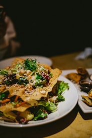 A plate of vibrant salad topped with crispy sesame crackers, mixed greens, and various vegetables is displayed. The dish is colorful and garnished with a variety of textures. It sits on a light-colored plate, accompanied by some rolls and a small bowl of sauce on the side.