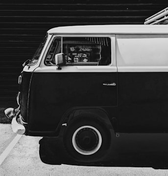 A classic van is parked against a dark background. The van has a two-tone paint job with a light upper part and a darker lower part. The image is in black and white, highlighting the contrast and texture. Various stickers are visible on the window, adding a personalized touch. The clean, reflective surface of the vehicle and the old-fashioned style give it a retro feel.