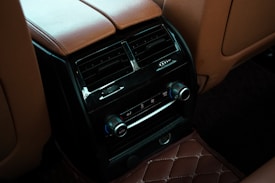 A close-up of the rear seat console in a car, featuring a dark glossy panel with air vents and climate control knobs. The surrounding area is upholstered in brown leather with visible stitching, and the floor mat has a quilted pattern.