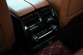 A close-up of the rear seat console in a car, featuring a dark glossy panel with air vents and climate control knobs. The surrounding area is upholstered in brown leather with visible stitching, and the floor mat has a quilted pattern.
