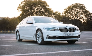 A sleek white car is parked in an empty parking lot. The backdrop consists of trees with sunlight filtering through, casting a warm glow. The car's design is modern with clean lines and a shiny exterior.
