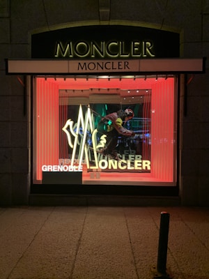 A storefront window display featuring the brand Moncler. The display includes illuminated signage with the word 'Moncler' in bold lettering and a stylized logo. Inside the window, there is a mannequin dressed in ski gear, including goggles and a helmet, holding ski poles. The background is vibrant with vertical red lights creating a striking contrast.