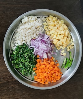 A round metal tray features neatly chopped vegetables, including cauliflower, potatoes, green beans, red onions, carrots, and green chili peppers. Each type of vegetable is organized into a distinct section, showcasing a variety of colors and textures.