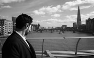 A man in a suit looks over a river from a bridge, observing the cityscape with a prominent skyscraper and historic buildings under a cloudy sky.