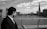 A man in a suit looks over a river from a bridge, observing the cityscape with a prominent skyscraper and historic buildings under a cloudy sky.