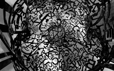 An intricate structure composed of various letters and symbols, forming a complex, layered pattern against the backdrop of a cloudy sky. The artwork appears three-dimensional and creates a sense of depth and texture.