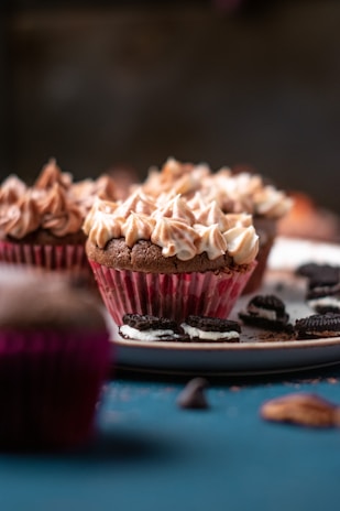 Cupcakes with swirled frosting sit on a plate. The cupcakes are in red paper liners and surrounded by broken pieces of chocolate cookies, which adds a decorative touch. The background has a soft focus, highlighting the cupcakes in the foreground.