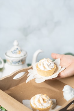 A hand holds a small dessert with white and light brown swirled cream on top, placed on a decorative cloth resembling petals. Nearby, there is a similar dessert in a brown cardboard tray along with a small white ceramic bird. In the background, a floral-patterned teapot with a matching lid can be seen, suggesting a delicate and elegant setting for tea or desserts.