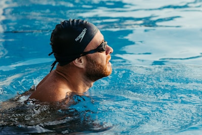 A person is swimming in a pool, wearing a black swim cap and goggles. The water around them is a clear blue, and sunlight creates reflections on the surface. The swimmer is focused, with a serene expression as they move through the water.