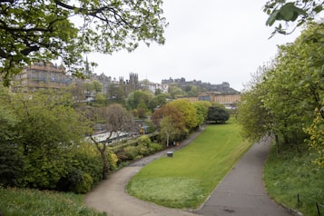 A lush green park with winding pathways surrounded by dense foliage and trees. In the distance, a cityscape featuring historic buildings and architectural structures is visible. A partly cloudy sky suggests it might be a slightly overcast day.