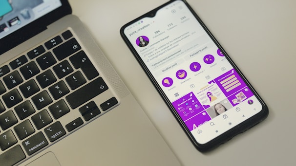 A smartphone displaying a social media profile screen with a bright purple theme, placed next to a laptop keyboard on a light-colored surface. The phone's screen shows various icons and text in French, along with profile details. The laptop's keyboard layout is visible, with black keys and white symbols.