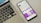 A smartphone displaying a social media profile screen with a bright purple theme, placed next to a laptop keyboard on a light-colored surface. The phone's screen shows various icons and text in French, along with profile details. The laptop's keyboard layout is visible, with black keys and white symbols.