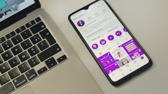 A smartphone displaying a social media profile screen with a bright purple theme, placed next to a laptop keyboard on a light-colored surface. The phone's screen shows various icons and text in French, along with profile details. The laptop's keyboard layout is visible, with black keys and white symbols.