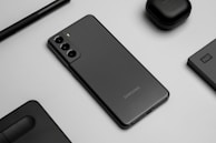 A flat lay image featuring a sleek black smartphone with three rear cameras, positioned on a gray surface. Surrounding the phone are matching black accessories, including wireless earbuds, a stylus pen, an external hard drive, and a laptop touchpad segment.