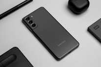 A flat lay image featuring a sleek black smartphone with three rear cameras, positioned on a gray surface. Surrounding the phone are matching black accessories, including wireless earbuds, a stylus pen, an external hard drive, and a laptop touchpad segment.