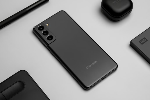 A flat lay image featuring a sleek black smartphone with three rear cameras, positioned on a gray surface. Surrounding the phone are matching black accessories, including wireless earbuds, a stylus pen, an external hard drive, and a laptop touchpad segment.