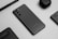 A flat lay image featuring a sleek black smartphone with three rear cameras, positioned on a gray surface. Surrounding the phone are matching black accessories, including wireless earbuds, a stylus pen, an external hard drive, and a laptop touchpad segment.
