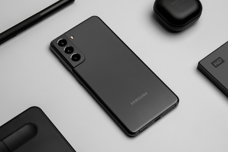 A flat lay image featuring a sleek black smartphone with three rear cameras, positioned on a gray surface. Surrounding the phone are matching black accessories, including wireless earbuds, a stylus pen, an external hard drive, and a laptop touchpad segment.