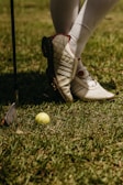 A person wearing white golf shoes and knee-high socks stands on grass. A yellow golf ball lies on the ground near a golf club with a metallic head.