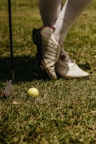 A person wearing white golf shoes and knee-high socks stands on grass. A yellow golf ball lies on the ground near a golf club with a metallic head.