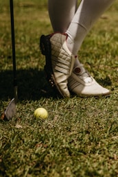 A person wearing white golf shoes and knee-high socks stands on grass. A yellow golf ball lies on the ground near a golf club with a metallic head.