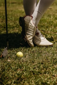 A person wearing white golf shoes and knee-high socks stands on grass. A yellow golf ball lies on the ground near a golf club with a metallic head.