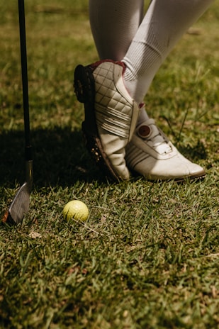 A person wearing white golf shoes and knee-high socks stands on grass. A yellow golf ball lies on the ground near a golf club with a metallic head.