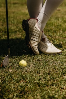 A person wearing white golf shoes and knee-high socks stands on grass. A yellow golf ball lies on the ground near a golf club with a metallic head.