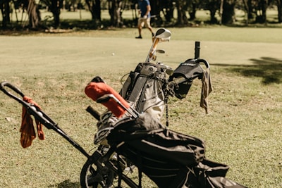 A golf bag filled with clubs is positioned on a grassy field, with a small trolley attached. Some bags have covers, and a towel is draped over one side. In the background, a person is walking across the green, surrounded by trees.