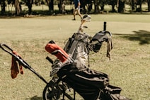 A golf bag filled with clubs is positioned on a grassy field, with a small trolley attached. Some bags have covers, and a towel is draped over one side. In the background, a person is walking across the green, surrounded by trees.