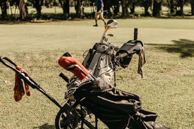 A golf bag filled with clubs is positioned on a grassy field, with a small trolley attached. Some bags have covers, and a towel is draped over one side. In the background, a person is walking across the green, surrounded by trees.