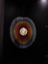 A spherical light fixture with a mosaic of colorful glass pieces hangs against a dark background. Surrounding the sphere is a circular pattern resembling a mandala, featuring a gradient of colors including red, orange, yellow, white, and blue. The intricate design gives a sense of symmetry and depth.