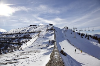 A snowy mountain landscape featuring a ski slope with several skiers and snowboarders. A line of ski lifts runs parallel to the slope, and snow fences line the hillside. The bright sun shines over the area, creating a clear sky with a few clouds for a bright, crisp scene.