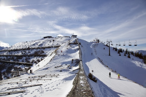 A snowy mountain landscape featuring a ski slope with several skiers and snowboarders. A line of ski lifts runs parallel to the slope, and snow fences line the hillside. The bright sun shines over the area, creating a clear sky with a few clouds for a bright, crisp scene.