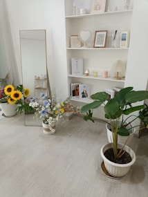 A modern interior space features a minimalistic white shelving unit displaying books, framed art, and decorative items such as a heart-shaped mirror. A tall mirror leans against the wall near a vibrant bouquet of mixed flowers including daisies and sunflowers. A large potted green plant is placed on the right side, adding a touch of nature to the decor. The light color palette and clean lines contribute to an airy and serene ambiance.
