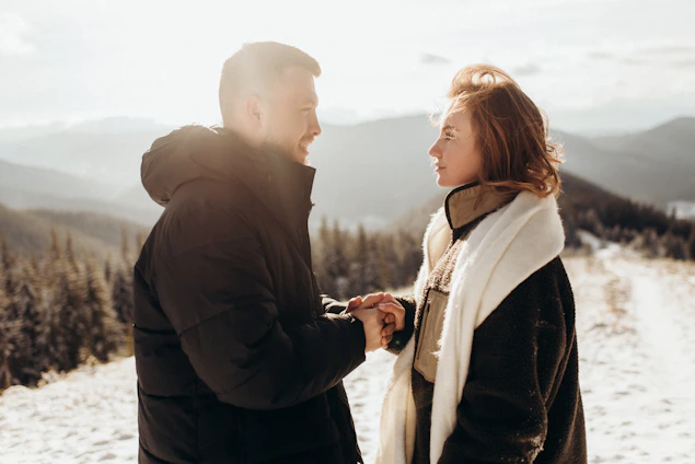 A cozy winter scene featuring a man and woman dressed in light-colored ski jackets and pants, standing against a snowy mountain backdrop.
