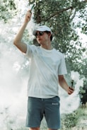 A person wearing a white cap and sunglasses is standing outdoors amidst greenery. They hold smoke emitters in both hands, with smoke surrounding them, creating a hazy atmosphere.