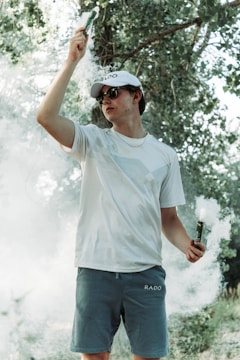 A person wearing a white cap and sunglasses is standing outdoors amidst greenery. They hold smoke emitters in both hands, with smoke surrounding them, creating a hazy atmosphere.
