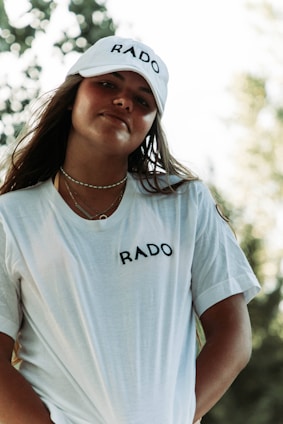 A person with long hair is wearing a white cap and t-shirt, both branded with the word 'RADO'. The background features blurred greenery and trees.