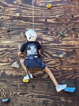 A child wearing a helmet and harness is climbing a wooden rock wall. The wall features various colored climbing holds, including yellow, green, and blue. The child is wearing a dark shirt, blue shorts, and white shoes.