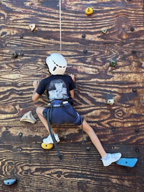 A child wearing a helmet and harness is climbing a wooden rock wall. The wall features various colored climbing holds, including yellow, green, and blue. The child is wearing a dark shirt, blue shorts, and white shoes.