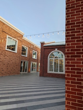 An outdoor courtyard with modern red-brick buildings featuring large windows and decorative string lights hung overhead. The paving is striped with light and dark gray tiles, creating a geometric pattern.