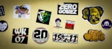 A collection of various stickers on a beige wall, including colorful designs and text. Some stickers have graphics of faces, logos, and text with phrases such as 'Zero Gomez' and 'Forza Sankt Pauli.' Other stickers include abstract designs, a skull and crossbones, and numbers like '20' and '07'.