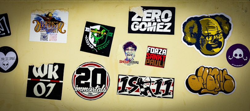 A collection of various stickers on a beige wall, including colorful designs and text. Some stickers have graphics of faces, logos, and text with phrases such as 'Zero Gomez' and 'Forza Sankt Pauli.' Other stickers include abstract designs, a skull and crossbones, and numbers like '20' and '07'.
