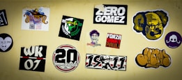 A collection of various stickers on a beige wall, including colorful designs and text. Some stickers have graphics of faces, logos, and text with phrases such as 'Zero Gomez' and 'Forza Sankt Pauli.' Other stickers include abstract designs, a skull and crossbones, and numbers like '20' and '07'.