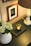 A warmly lit, cozy setting features a small lamp with a beige shade on a dark surface. Next to the lamp is a green candle placed on top of a closed book, creating a soothing and intimate atmosphere. In the foreground, a white vase holds pale hydrangea flowers. Additionally, there are framed pictures or artwork on the wall, adding an elegant touch to the scene.