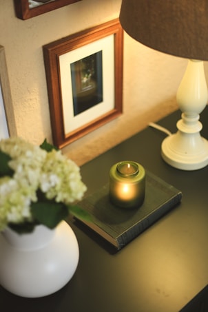 A warmly lit, cozy setting features a small lamp with a beige shade on a dark surface. Next to the lamp is a green candle placed on top of a closed book, creating a soothing and intimate atmosphere. In the foreground, a white vase holds pale hydrangea flowers. Additionally, there are framed pictures or artwork on the wall, adding an elegant touch to the scene.