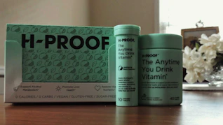 A collection of H-PROOF products including a box and two containers labeled as 'The Anytime You Drink Vitamin.' The packaging highlights health benefits such as supporting alcohol metabolism and promoting liver health. The products are vegan, gluten-free, and sugar-free. They are placed on a wooden surface with a vase of white flowers in the background.