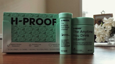 A collection of H-PROOF products including a box and two containers labeled as 'The Anytime You Drink Vitamin.' The packaging highlights health benefits such as supporting alcohol metabolism and promoting liver health. The products are vegan, gluten-free, and sugar-free. They are placed on a wooden surface with a vase of white flowers in the background.